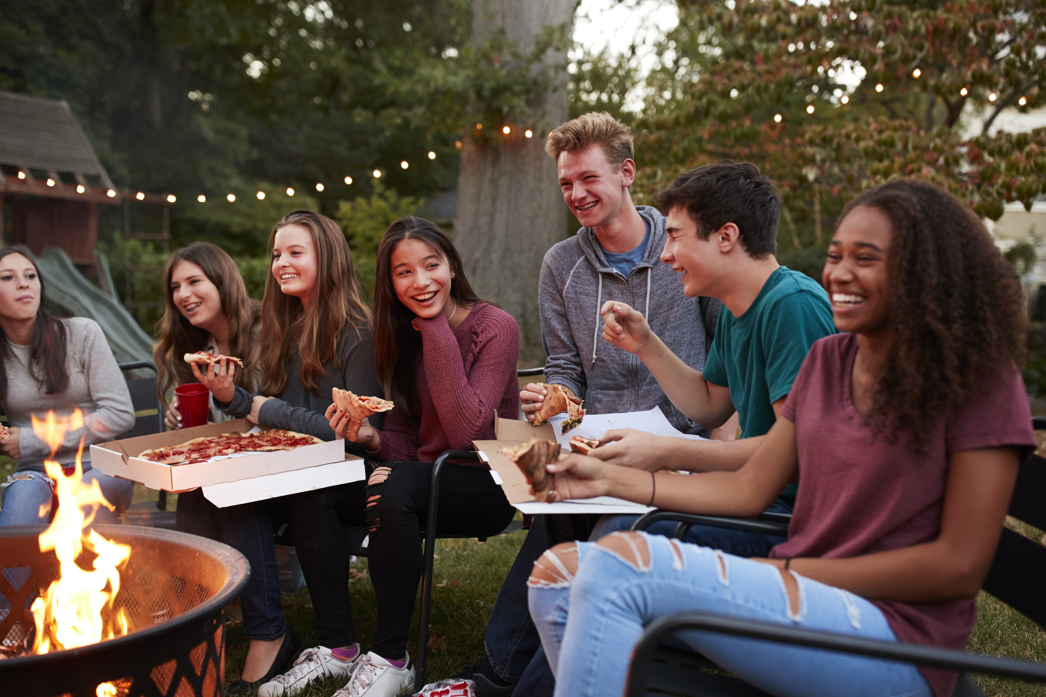 Seven teens gathered around a fire pit while eating delivery pizza
