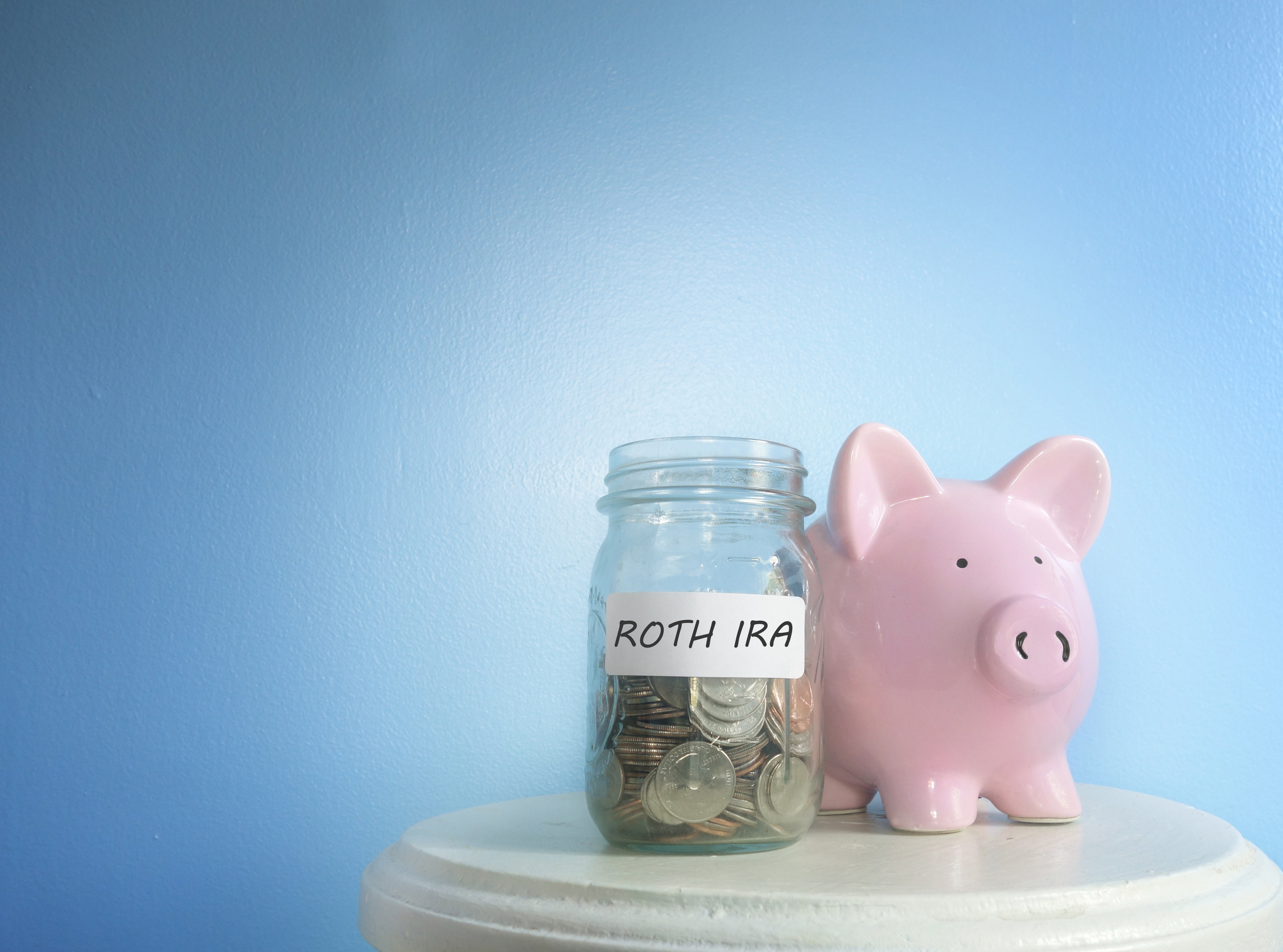 A jar with the label ROTH IRA filled with coins next to a pink piggy bank on a little round table.