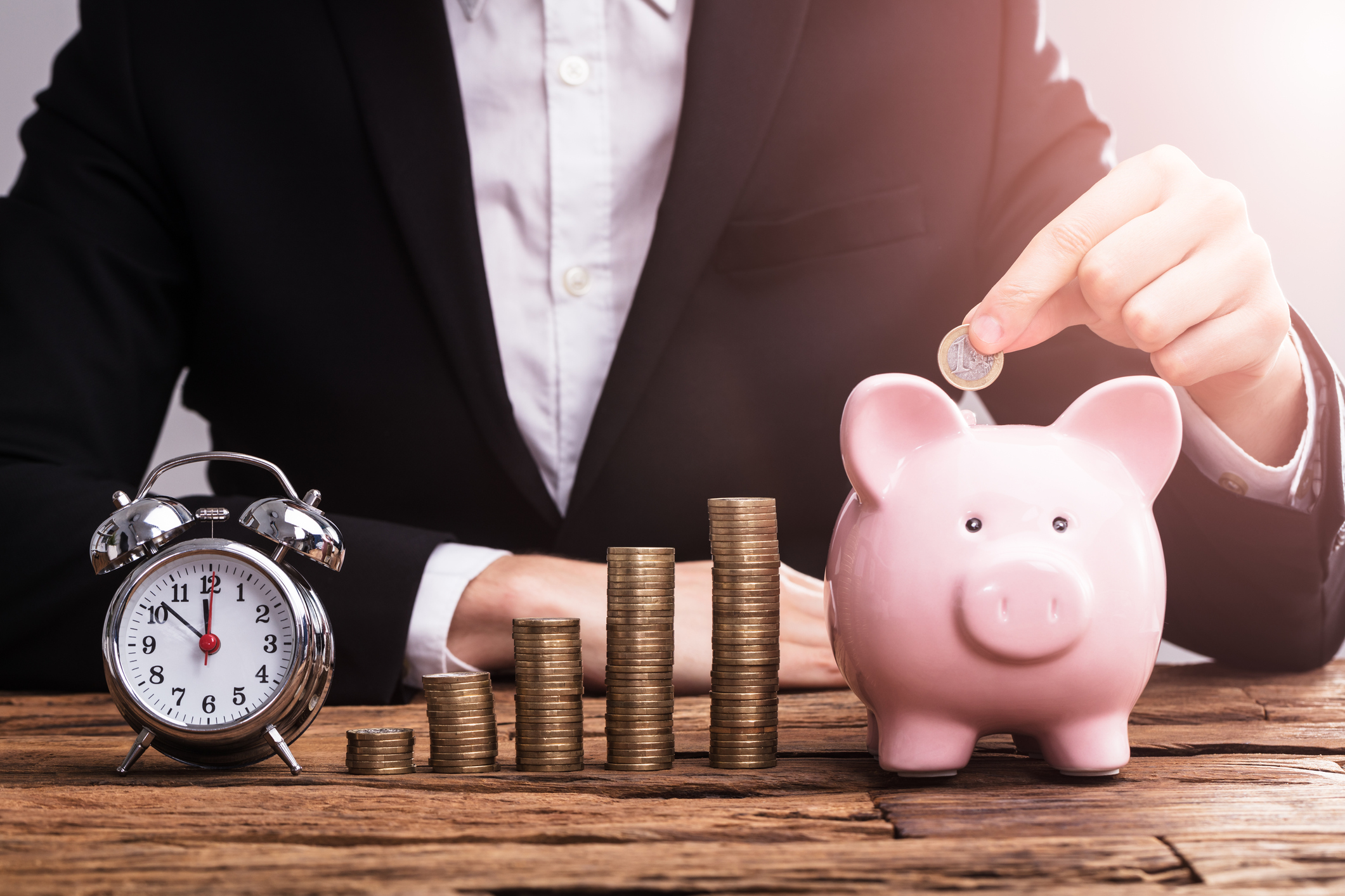 With an alarm clock and growing stacks of coins in front of him, a man drops a coin into a piggy bank.