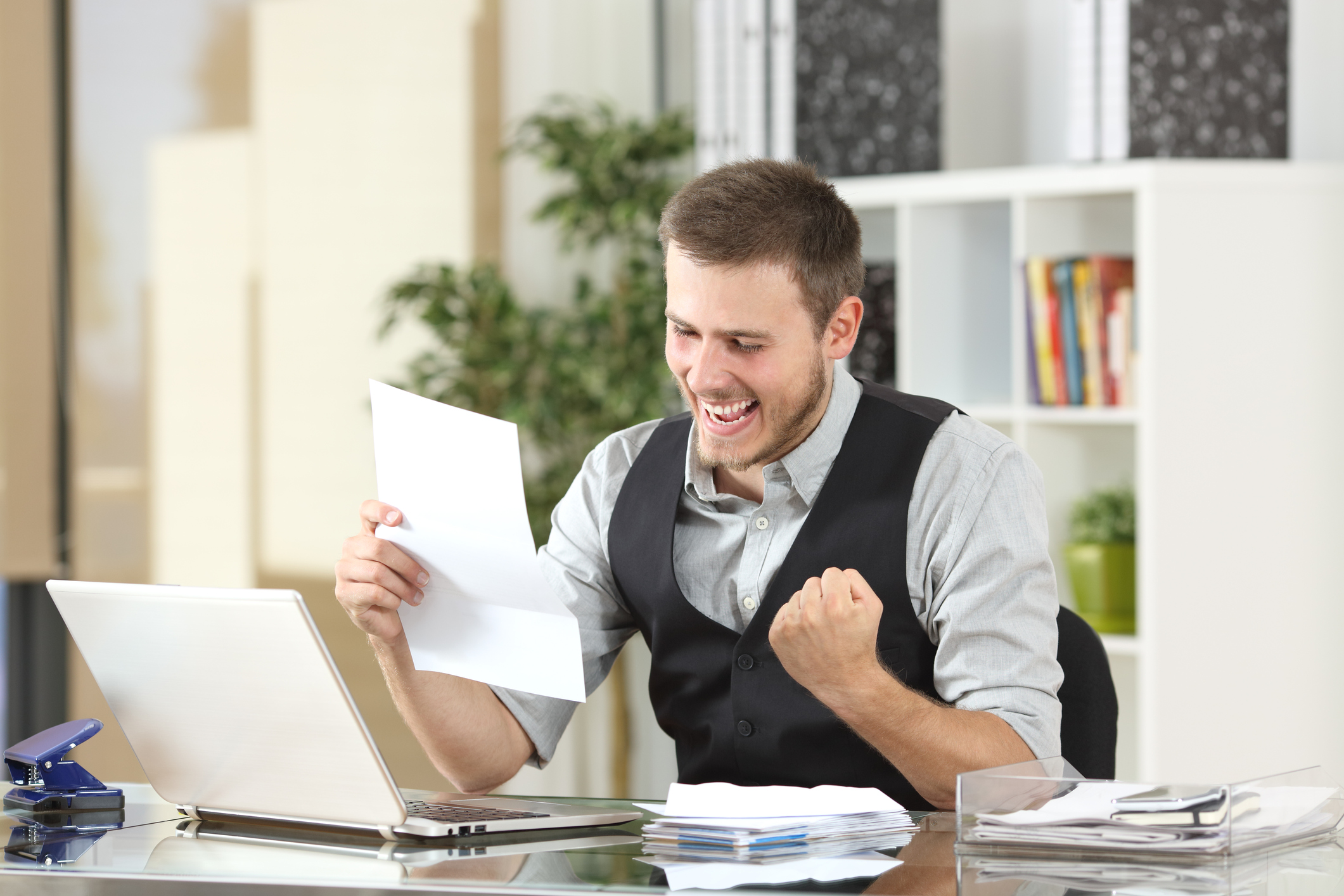 A man at a desk with a laptop, holding a piece of paper up and celebrating.