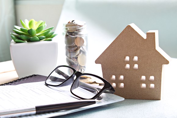 Miniature house next to a pair of glasses, a pen, a small jar filled with coins, and a small succulent in a pot