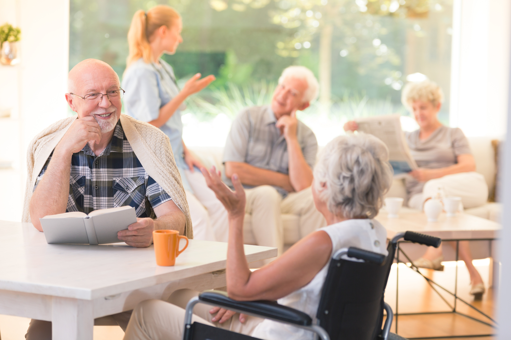 Elderly man talking with woman in wheelchair while sitting together at table in common room in senior living facility