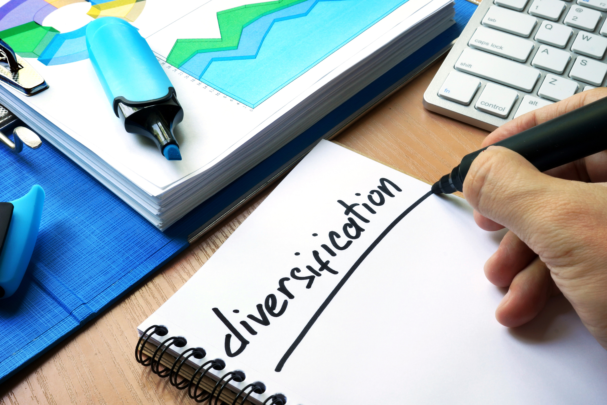 Hand writing "diversification" on a notebook page next a paper showing stock charts, a highlighter, and a computer keyboard