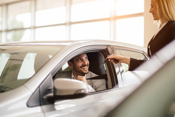 Woman handing car keys to man sitting in driver's seat.