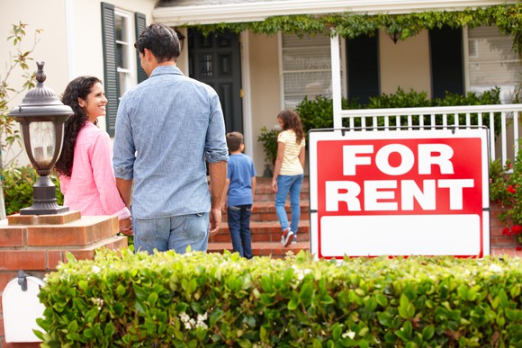 Family of four standing outside house with a For Rent sign