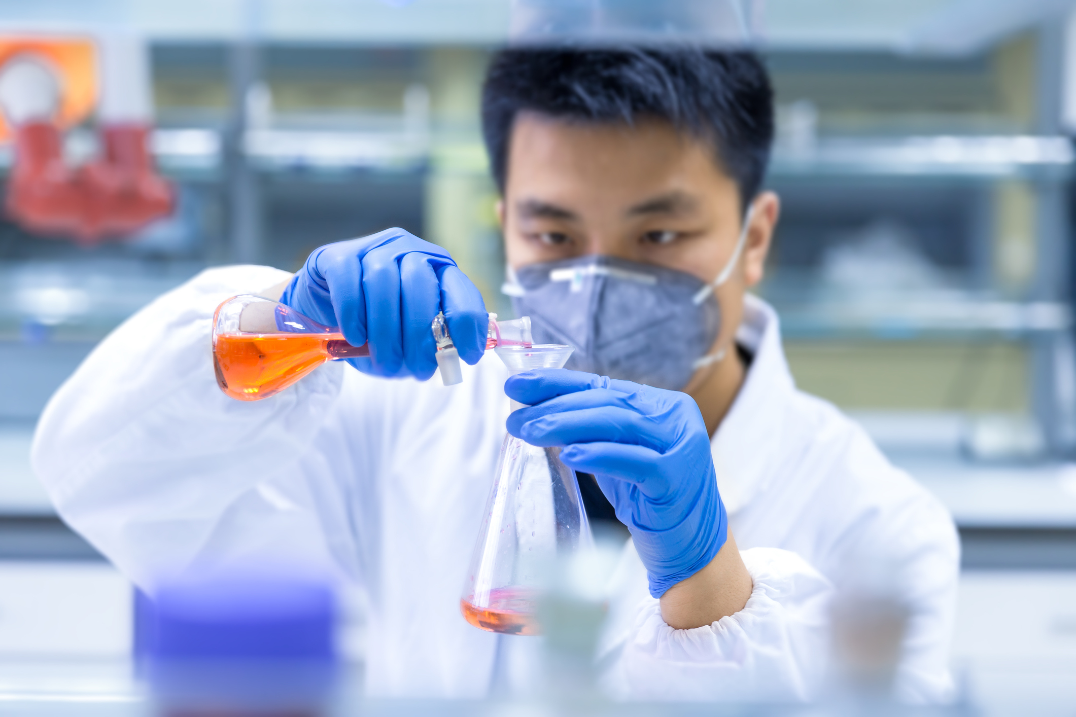 A scientist wearing a mask pours liquid from one flask into another