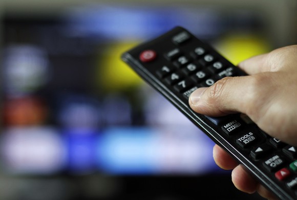 Hand holding a remote control in the foreground; blurred screen in background