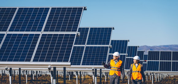 Two workers in orange vests and white hard hats walk past a field of solar panels.