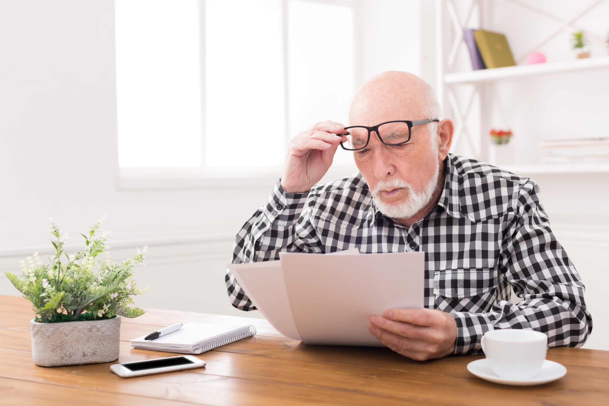 Senior man with his glasses raised slightly reading important documents while sitting at a desk with a plant, an iphone, a paid, and a coffee cup.