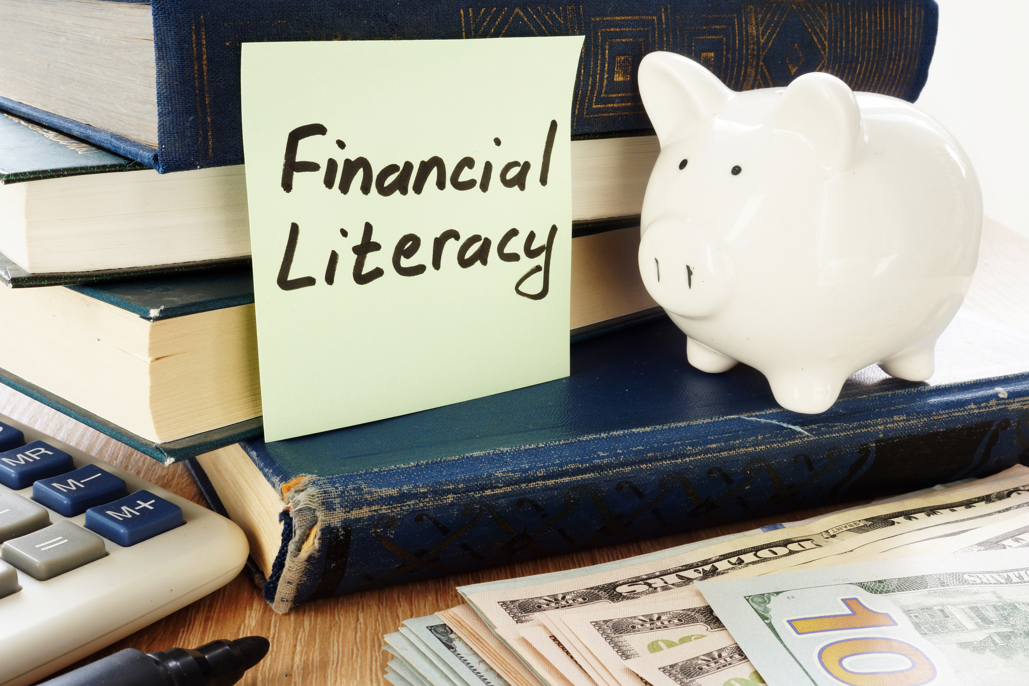 Stack of books next to a piggy bank, cash, a calculator, and a sticky note that says "Financial Literacy"
