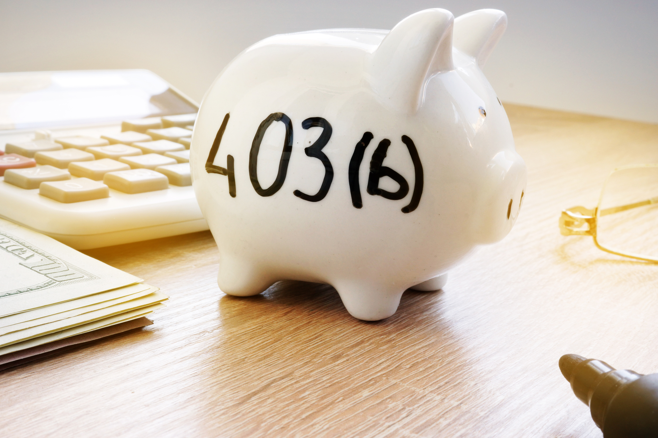A piggy bank labelled "403(b)" sits on a desk near a stack of currency and a calculator.