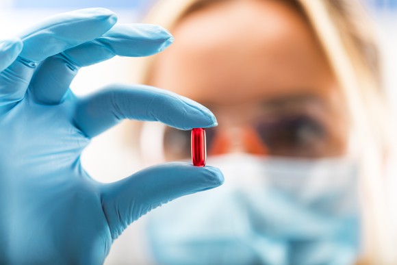 Woman wearing surgical mask examining a red pill in her gloved hand