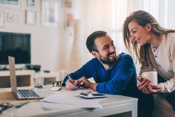 Man at a desk smiling at woman behind him who is holding a mug and looking at what is on his computer screen.