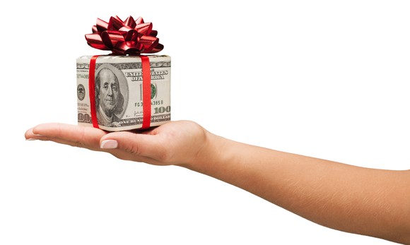 An outstretched arm and hand holding a box made of $100 bills with a bow on top.
