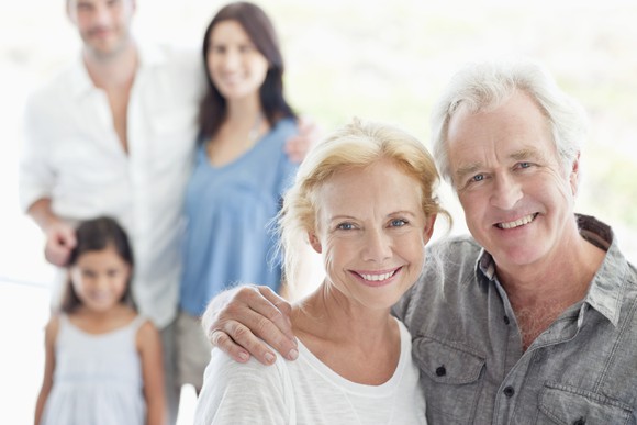 A mature couple standing together with the husbands arm around the wife's shoulder. A young family stands in the background.