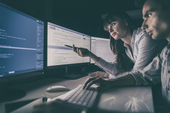 Two people working on a computer with three monitors.
