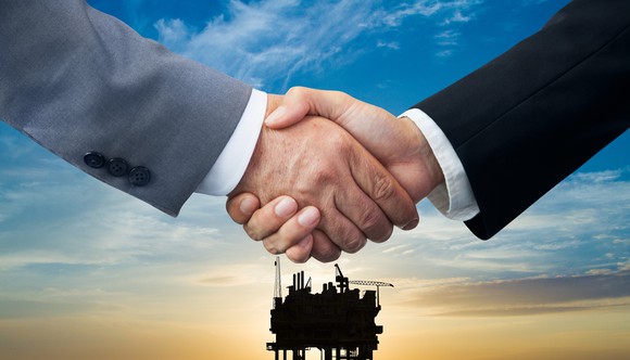 Two men shake hands with an offshore oil rig in the background.