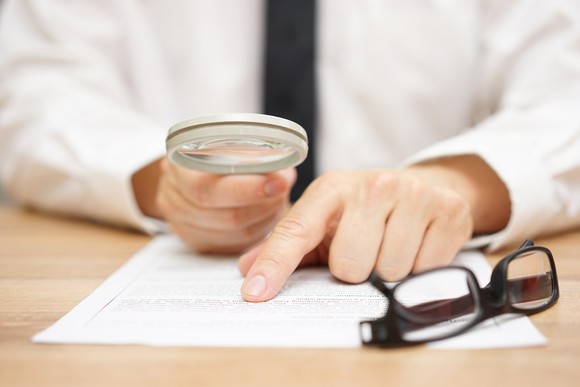 A hand holding a magnifying glass over a piece of paper, next to a folded pair of glasses.
