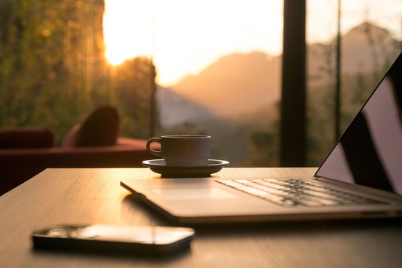 Laptop and coffee mug sitting on desk in front of window with view of sunrise coming over mountains