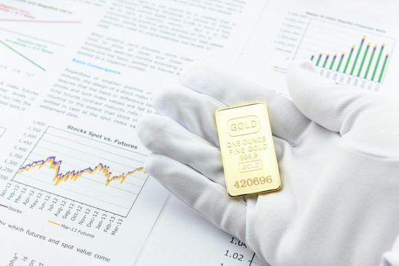 A person with a white glove holding a gold ingot over a rising stock chart.