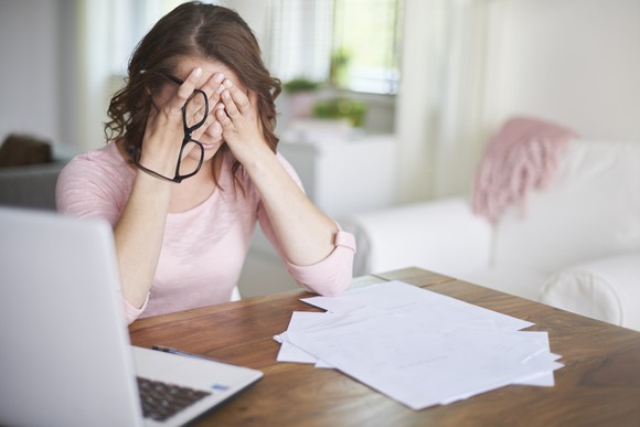 Woman sitting at a table holding her glasses with her eyes closed, stressing out over finances