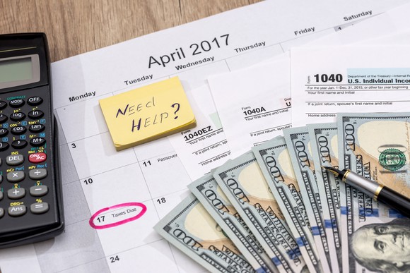 Tax forms, cash, a pen, and a calculator sitting on a calendar with Tax Day circled, and a sticky note with the words "need help?"