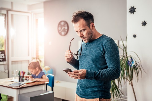 A father looks at his wireless phone with his daughter in the background.
