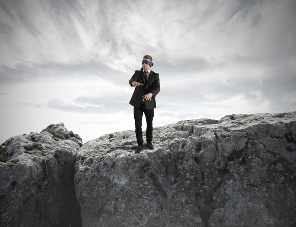 A man blindfolded and walking perilously on rocks.