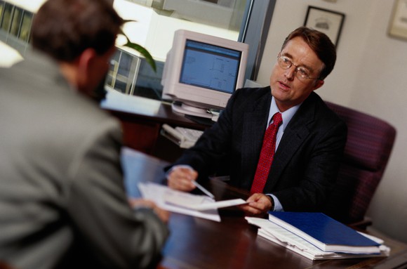 A man in a suit consulting another, at a desk.