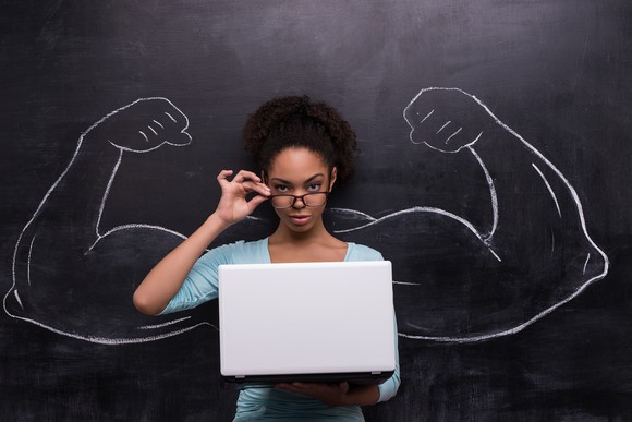 Young African-American woman with laptop against chalkboard background. Two strong muscular arms drawn on chalkboard
