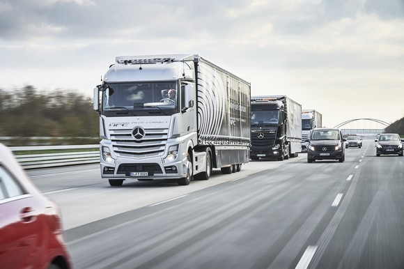Three Mercedes-Benz tractor-trailers are shown on a highway. The driver of the lead truck has his hands behind his head, not touching the steering wheel.