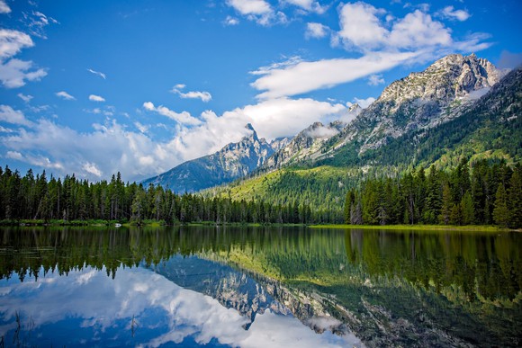 String Lake's glassy surface reflects the surrounding evergreens and mountains, along with the clear blue sky