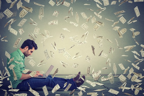 A man sitting with his laptop while cash showers down on him.