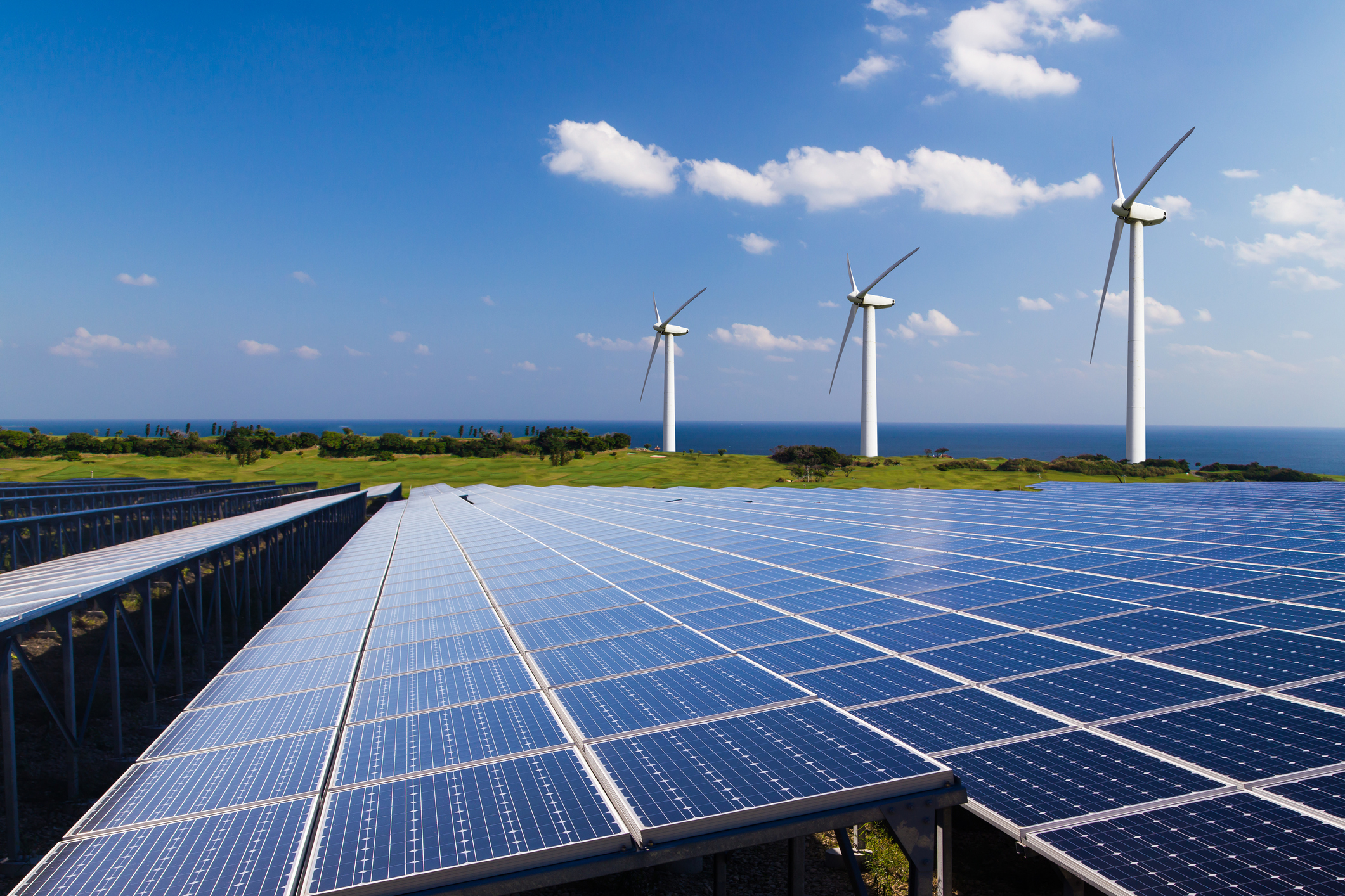 A solar-panel array, with wind turbines in the background against a blue sky.