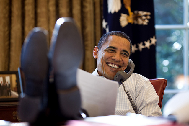 Former President Barack Obama smiling with his feet up on his desk in the Oval Office.