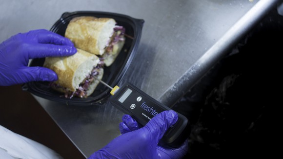 A restaurant worker wearing blue vinyl gloves inserts a Fresh Temp connected thermometer into a sandwich.