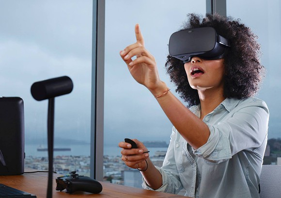 A woman sits at a desk wearing an Oculus virtual reality headset, pointing up at something she must be seeing inside the virtual world.