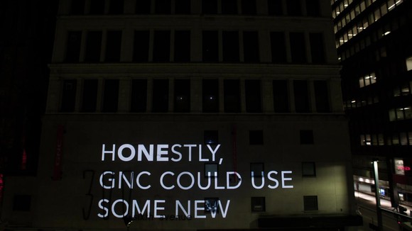 Video image message projected on side of building, saying "Honestly, GNC could use some new"