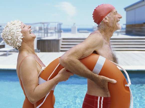 An elderly man and woman next to a swimming pool smile as they walk by.