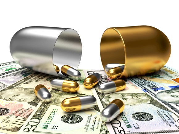 Silver and gold pills spill out of a larger silver and gold pill onto a pile of money.
