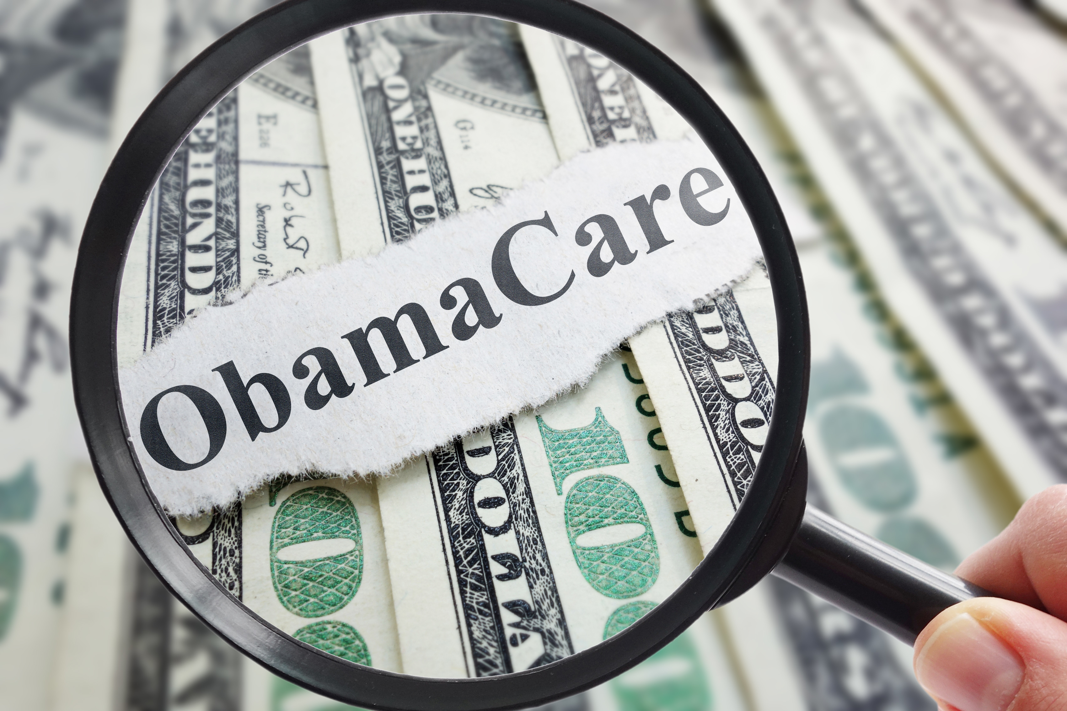 Magnifying glass looking at word "Obamacare" set against dollar bills