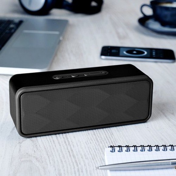 A speaker on a tabletop, with other items including a laptop, a cup of coffee, a pen, and paper.