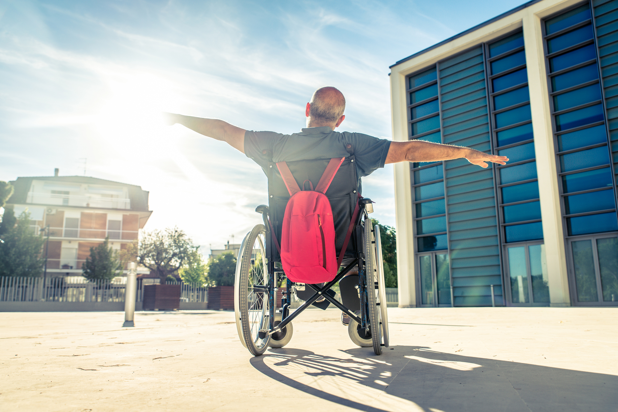 A man in a wheelchair outside of a building raises his arms vertically as if to fly.