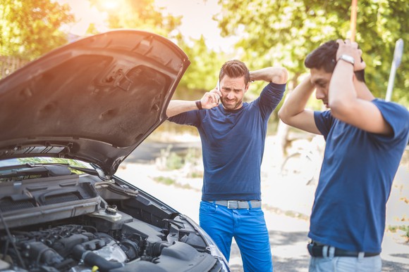 Two men stand next to broken-down car with open hood.