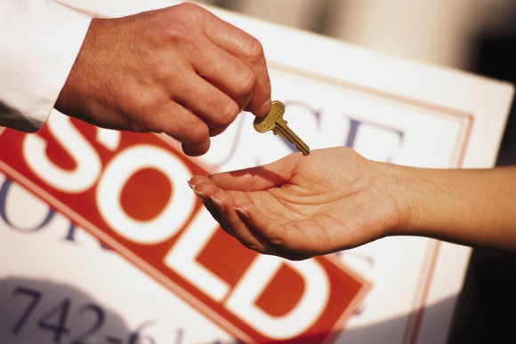 Hand receiving a new home key with "Sold" sign in the background.