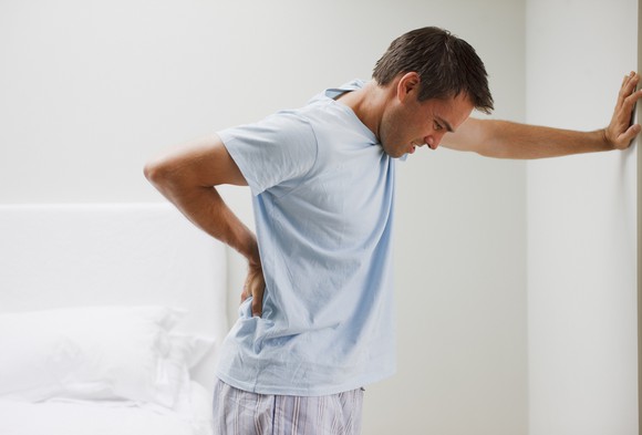 A man leans against a wall holding his back in pain.