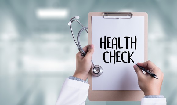 Two white-sleeved arms, holding a stethoscope, a pen, and a paper reads "health check" attached to a clipboard.