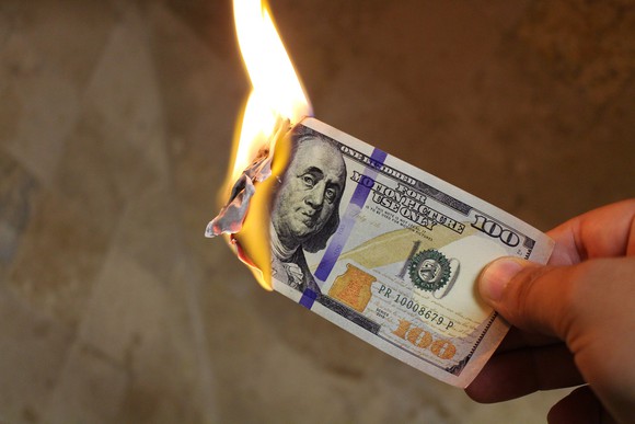 A hand holds a burning $100 bill.