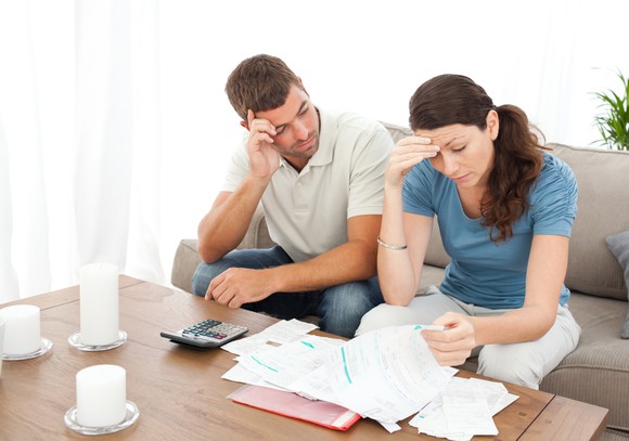 A couple sits at a table, reviewing their pile of bills and looking stressed.