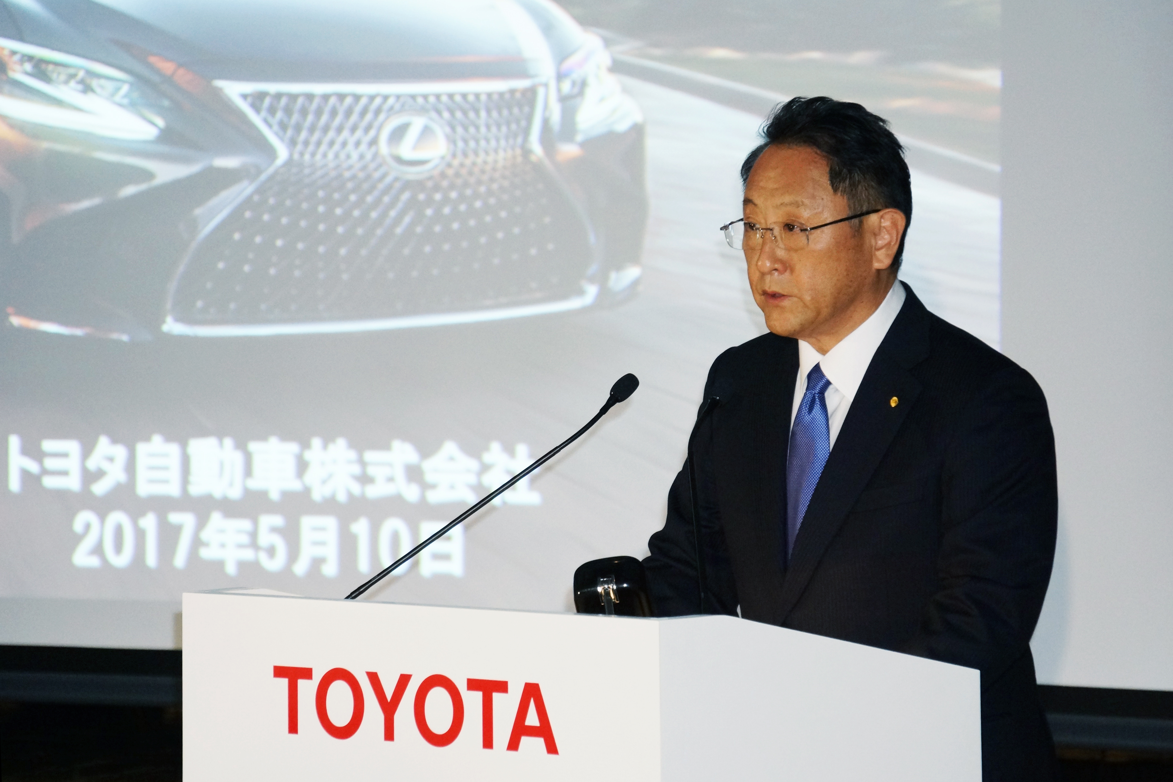 Toyoda is shown speaking at a podium in front of a slide screen.